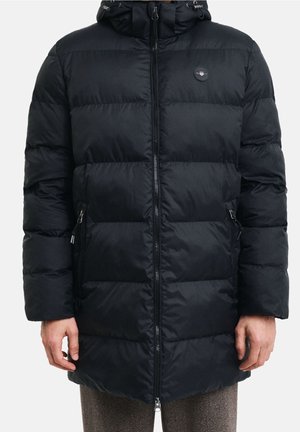 Schwarze gesteppte lange Steppjacke mit Frontreißverschluss, seitlichen Reißverschlusstaschen und Kapuze, getragen über brauner Hose an einer stehenden Person.