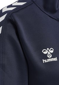 Granatowa bluza sportowa z gładkiego materiału, z białym logo Hummel i kontrastowym białym wzorem chevron na ramieniu.