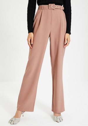 Vrouw draagt hoge taille, wijde broekspijpen in stoffig roze met een bijpassende riem, zwart topje met lange mouwen en gestreepte hakken met strik.