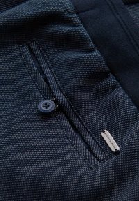 Tessuto blu scuro con un sottile motivo geometrico, dotato di una tasca con cerniera e di un grande bottone nero, insieme a un'etichetta metallica del marchio.