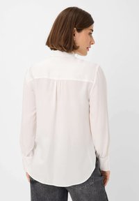 Blusa bianca a maniche lunghe con colletto alto, realizzata in tessuto leggero, con schiena plissettata e orlo arrotondato. Indossata con jeans scuri.