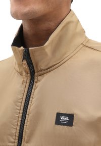 Beige Zip-Jacke mit hohem Kragen, ausgestattet mit einem schwarzen Reißverschluss und einem schwarzen Vans-Logo-Patch auf der Vorderseite. Glatte Stofftextur.