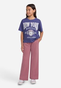 Ljubičasta pamučna majica sa grafikom "NEW YORK", uparen sa ružičastim hlačama širokih nogavica. Bele patike upotpunjuju odjevnu kombinaciju.
