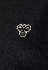 Svart tyg med en närbild av en vit broderad bi-logotyp, som har detaljerade svarta accenter och struktur i sömmen.