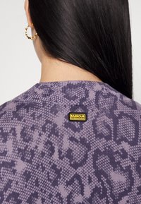 Gros plan d'une personne aux longs cheveux noirs portant un haut violet à motifs avec un écusson du logo Barbour International sur le haut du dos et une boucle d'oreille en anneau doré.