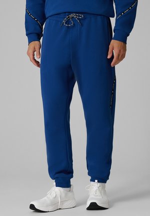 Homme portant un pantalon de survêtement bleu avec un cordon à la taille et des chaussures de sport blanches sur un fond gris.