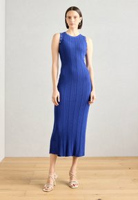 Robe sans manches bleu royal en maille côtelée avec détails en métal sur les épaules, design ajusté et longueur midi. Associée à des talons neutres à lanières.