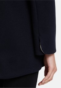 Marineblauer Wollmantel mit strukturierter Oberfläche und leicht ausgestellten Ärmeln. Ein weißes Innenfutter ist am Bund sichtbar.