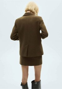 Personne blonde debout, vue de dos, portant un blazer marron et une jupe avec des bottes noires montantes jusqu'aux genoux, sur un fond uni.