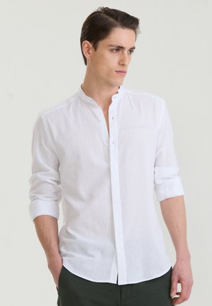 Jeune homme portant une chemise blanche à col mandarin avec les manches retroussées, regardant sur le côté sur un fond uni.