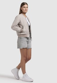 Lichtbeige bomberjack met zwarte rits, ribbels aan de mouwen en zijzakken, gecombineerd met grijze denim shorts en witte hoge sneakers.