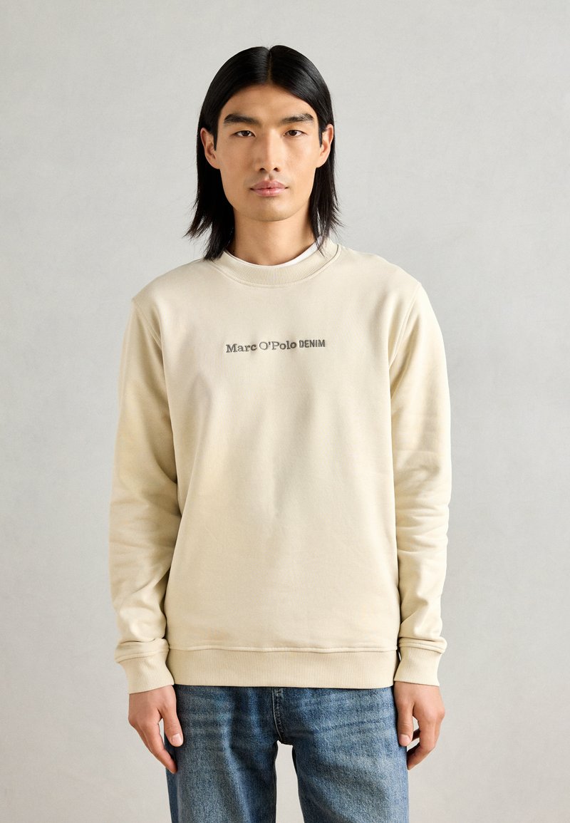 Šviesiai rudas džemperis, pagamintas iš minkštos medžiagos, su apvalia iškirpte ir ilgomis rankovėmis. Tamsiai pilka "Marc O’Polo DENIM" logotipas atspausdintas priekyje.