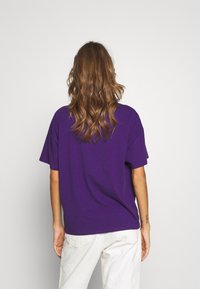 Lila bomull t-shirt med kort ärm och avslappnad passform, i kombination med benvita byxor. Visad bakifrån med vågigt hår.