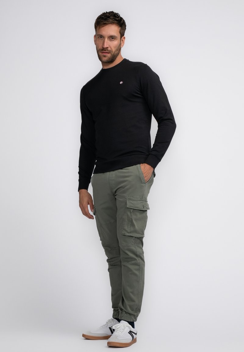 Zwarte longsleeve shirt, olijfgroene cargobroek met zijzakken, witte sneakers met zwarte accenten, staande tegen een effen achtergrond.