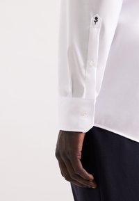 Camicia bianca a maniche lunghe con polsino abbottonato, caratterizzata da un ricamo floreale nero sulla manica. Tessuto morbido, vestibilità sagomata.