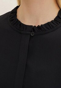 Blusa nera con colletto a volant, chiusura con bottoni e texture di tessuto liscia. Il design è semplice e su misura.