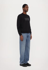 Felpa nera con un patch rettangolare, abbinata a jeans blu larghi e sneaker nere. Tessuto morbido; vestibilità casual.