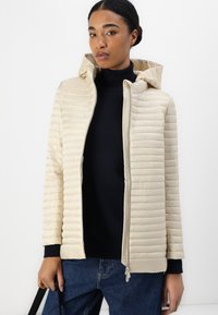Beige gewatteerde pufferjas met horizontaal doorgestikt ontwerp, ritssluiting aan de voorkant en ribgebreide manchetten. In combinatie met een zwarte turtleneck en denim.