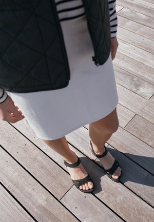 Person iført hvid nederdel, stribet top, sort foret vest og sorte sandaler står på trædæk med sollys og skygger.