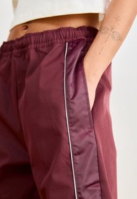 Burgundské sportovní kalhoty s hladkým povrchem, s bílým bočním pruhem a elastickým pasem. Jsou viditelné boční kapsy.