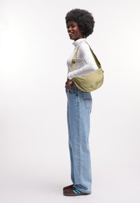 Bolso tipo bandolera verde de tela con correa ajustable, forma redondeada, llevado sobre el hombro; modelo con jersey blanco y jeans azules.