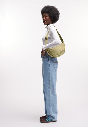Borsa a tracolla in tessuto verde con tracolla regolabile, forma arrotondata, indossata a spalla; modello con dolcevita bianca e jeans blu.