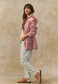 Rosa wasserdichte Jacke mit Kapuze, elastischem Bund und Taschen, kombiniert mit hellblauen Hosen und braunen Sneakern mit weißen Details.