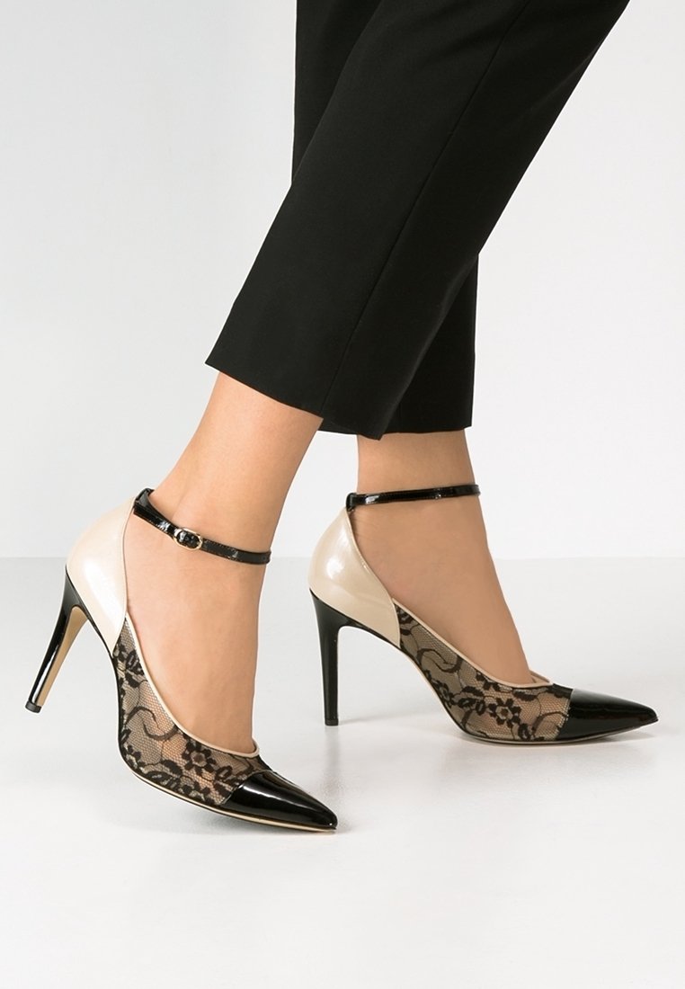 Femme portant des chaussures à talons hauts noires et beiges avec un détail en dentelle et des brides à la cheville, associées à un pantalon noir raccourci, debout sur un sol blanc.