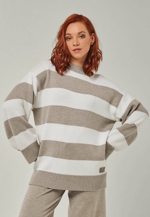 Frau mit roten Haaren, die einen beige-weiß breit gestreiften Pullover und beige Hose trägt, steht mit den Händen in der Hüfte vor einem einfarbigen Hintergrund.