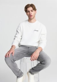 Felpa bianca con collo tondo e logo sul petto, abbinata a jeans grigi chiari, indossata con sneakers bianche, seduto su una sedia trasparente.