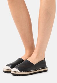 Nohy ve černých kožených espadrilách na slip-on s bílými švy z juty na jednobarevném světlém pozadí.