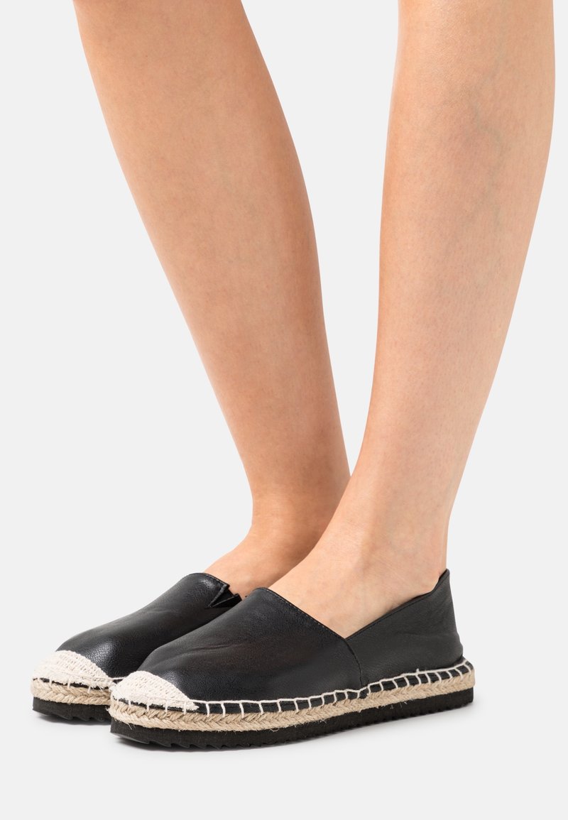 Nohy ve černých kožených espadrilách na slip-on s bílými švy z juty na jednobarevném světlém pozadí.