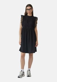 Robe noire avec un corsage en dentelle, manches courtes à volants, taille élastique et jupe fluide. Assortie avec des baskets de sport et des chaussettes blanches.