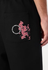 Black pants featuring a back pocket with a pink cherry blossom graphic and circular text, crafted from soft, stretchy fabric.