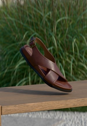 Sandale en cuir marron avec un design à brides croisées, détail de boucle à la cheville et une semelle en caoutchouc noir texturée.