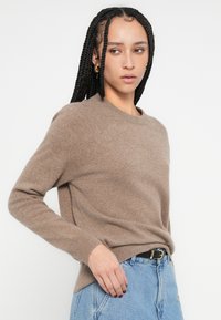 Καφέ πλεκτό πουλόβερ με στρογγυλή λαιμόκοψη, μανίκια ράγκλαν και απαλή υφή, σε συνδυασμό με ανοιχτόχρωμο τζιν παντελόνι.