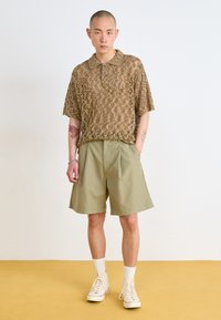Beige, teksturert kortermet polo med krage og knappelukking, sammen med khaki skjorteshortser med legg. Hvite høyhælte sneakers fullfører antrekket.