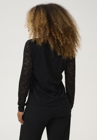 Top lungo a maniche nere in pizzo con un motivo floreale e superficie testurizzata. Retro liscia con design aderente, abbinato a pantaloni neri.