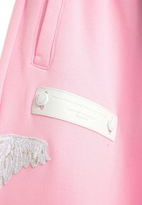 Tela rosa con una superficie texturizada, que presenta una etiqueta blanca con broches a presión y un diseño de alas bordado en blanco y dorado.