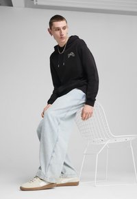 Felpa nera con cappuccio e logo, jeans a gamba larga blu chiaro e sneakers beige con suole in gomma, seduto su una sedia bianca in metallo.