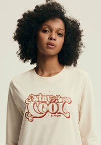 Jasno-beżowa koszula z długim rękawem z czerwonym i złotym retro napisem "stay COOL Be Happy". Wykonana z miękkiej bawełny, o luźnym kroju.