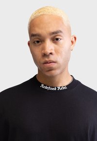 Czarna bluza z okrągłym dekoltem z napisem "Redefined Rebel" w białym kolorze. Model ma jasnoblond włosy i krótko przystrzyżoną fryzurę.