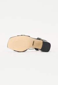 Zwarte sandaal met hakken, voorzien van een glad oppervlak, open teen en enkelbandje. Met een rubberen loopzool met ribbels en een beige binnenzool. Merklogo zichtbaar.