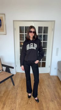 Žena oblečená v čiernej mikine GAP, čiernych nohaviciach a čiernych topánkach stojaca v interiéri na drevenej podlahe pred dvojitými dvernami so sklom.
