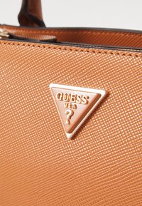 Ruskea teksturoitu käsilaukku, jossa on kullanvärinen kolmion muotoinen logolevy, jossa lukee "GUESS USA". Laukussa on vetoketjusuljenta ja sileä nahkareunus.