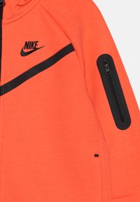 Oranžová mikina Nike so čiernym šikmým pruhom, čiernou zipsovou kapsou na rukáve a čiernym logom Nike na hrudi.