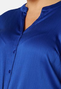 Chemise en satin bleu avec un motif texturé subtil, dotée d'un col mao et d'une fermeture à un bouton au niveau de l'encolure.