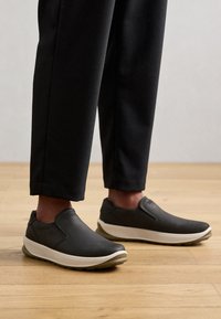 Chaussures à enfiler en cuir gris foncé avec une texture lisse, semelle intermédiaire blanche et semelle extérieure en caoutchouc flexible, accompagnées de pantalons noirs fuselés.