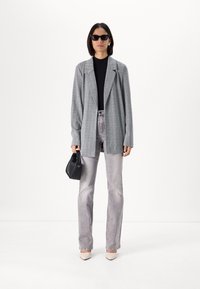 Gray plaid blazer with long sleeves, paired with a black top and light gray jeans. Holds a small black bag, wearing dark sunglasses.