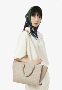 Borsa a tracolla beige con un motivo geometrico in rilievo, dotata di due manici superiori e di una tracolla removibile, esposta su uno sfondo semplice.